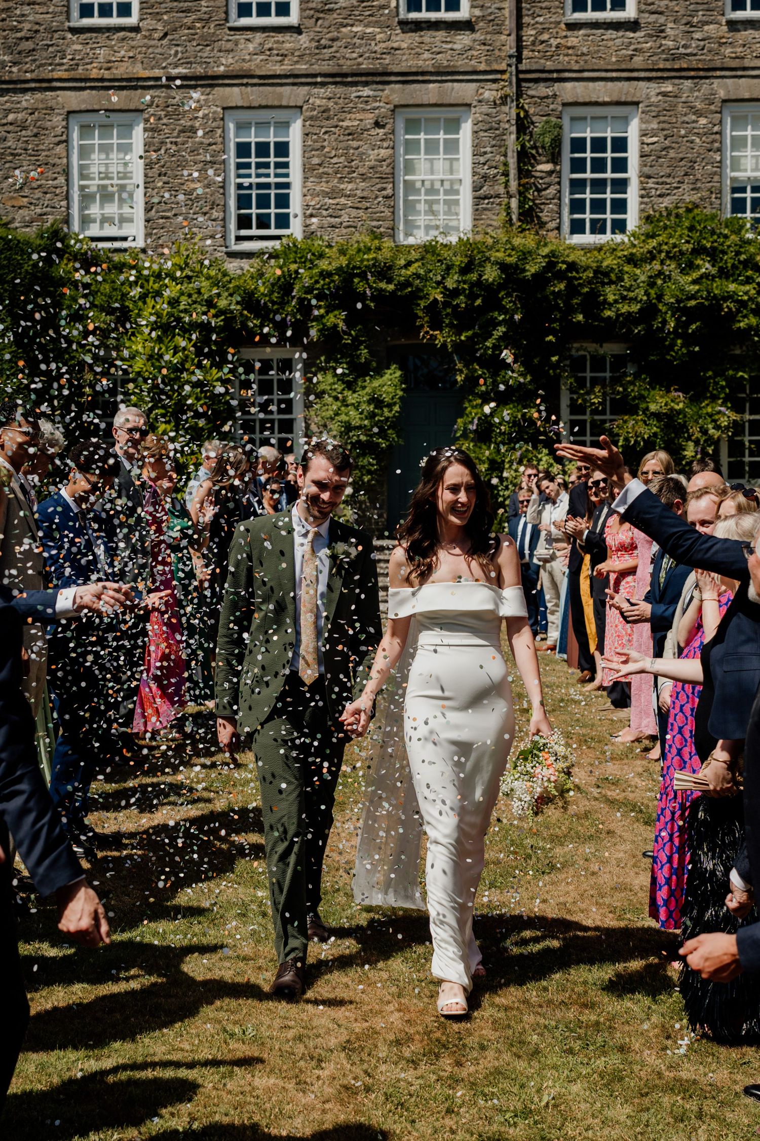 Bride and groom walk through confetti shower outside stone manor house surrounded by cheering wedding guests on sunny day.