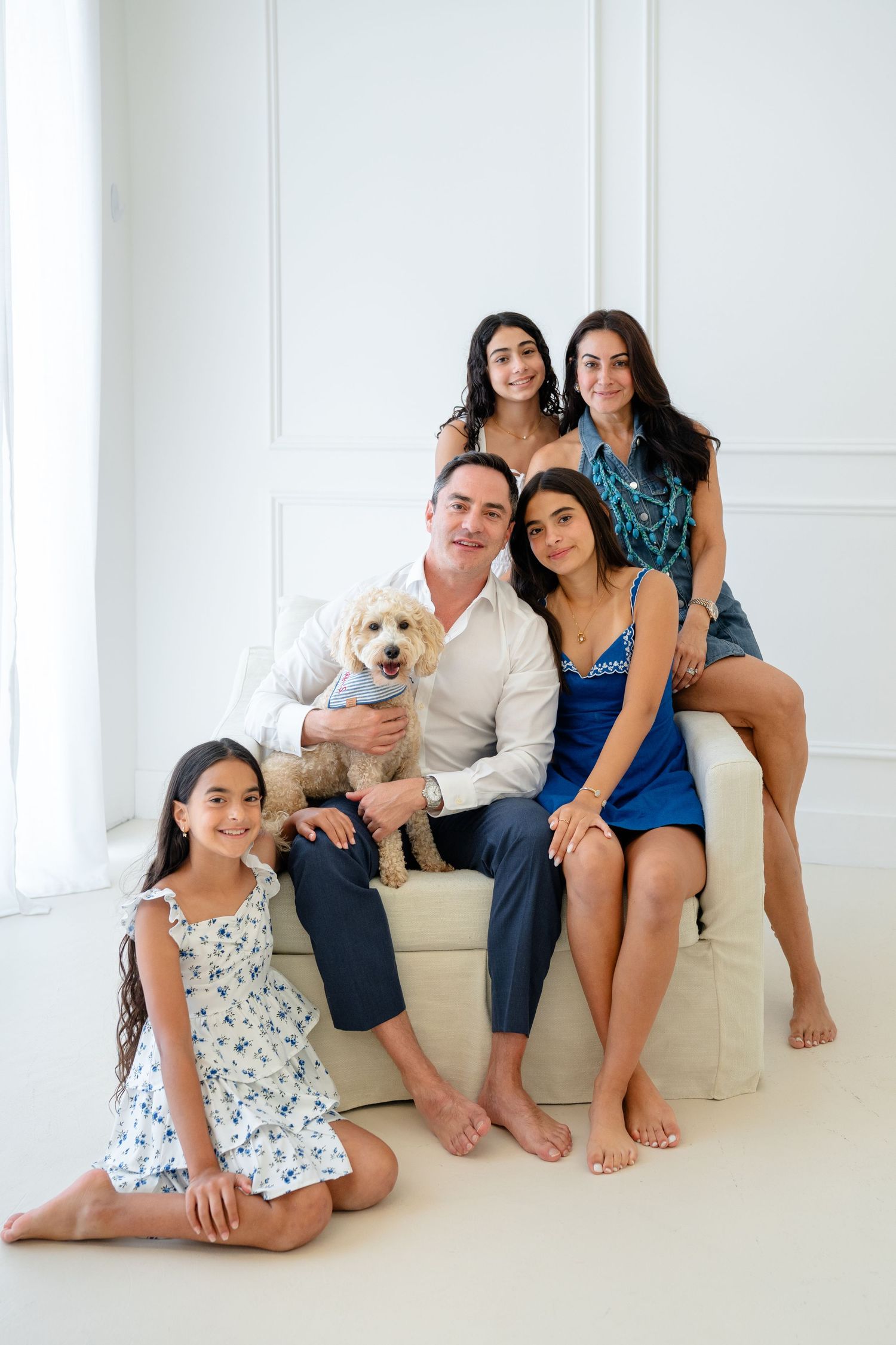 A happy family of five poses with their fluffy dog on a white sofa in a bright studio setting.