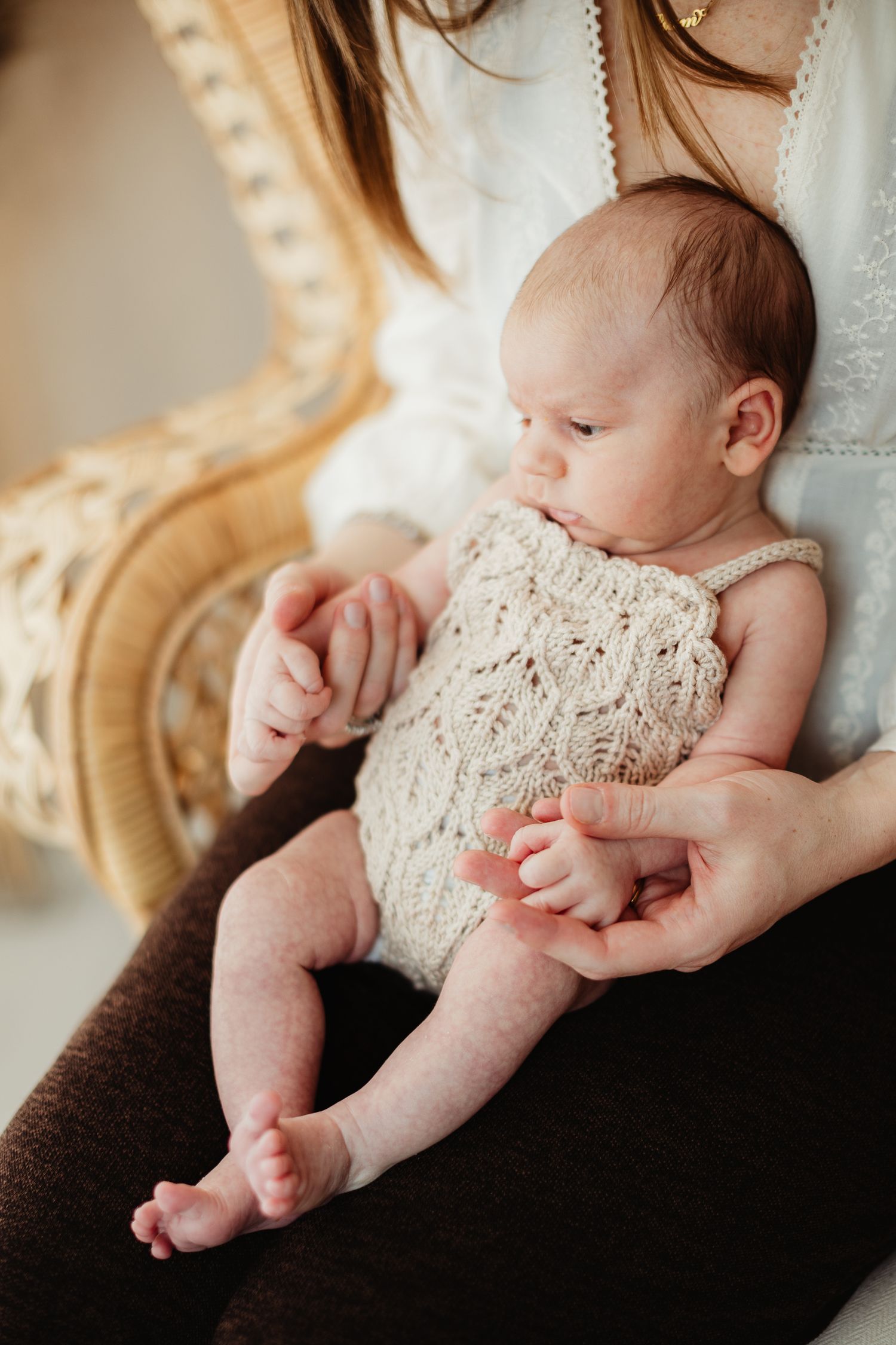 Newborn baby in knit romper resting on mother's lap in a wicker chair, with tiny hands and bare feet visible.