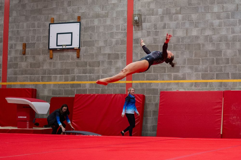 Gymnast performing a split leap on red tumbling mats in a gymnasium with a basketball hoop in background.
