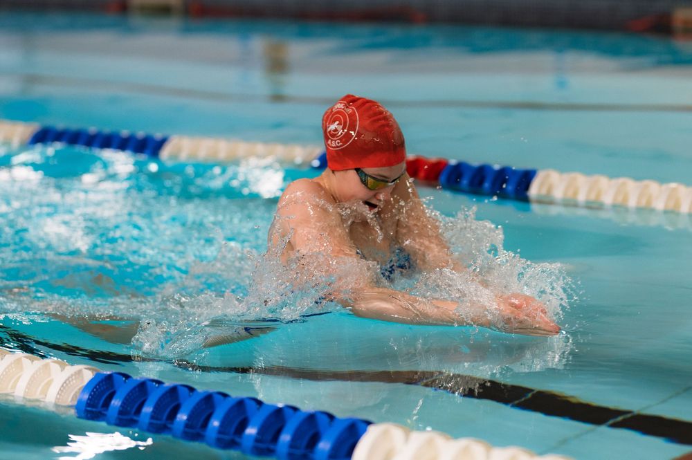 Swimmer in red cap performs breaststroke during competition with splashing water and blue lane dividers.