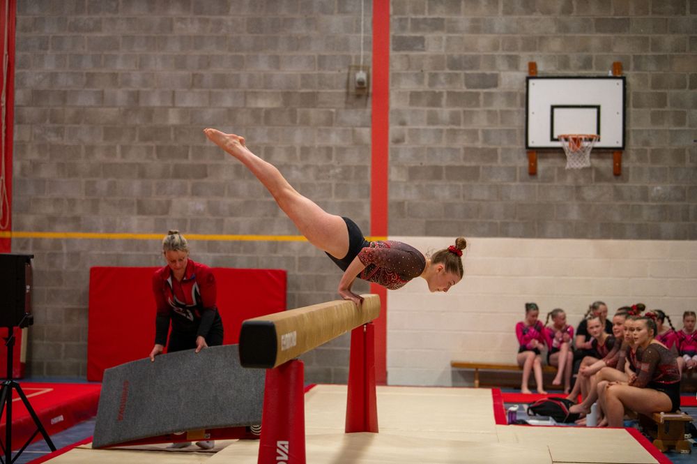 Gymnast performs a horizontal split on balance beam during competition while spectators watch from sideline mats.