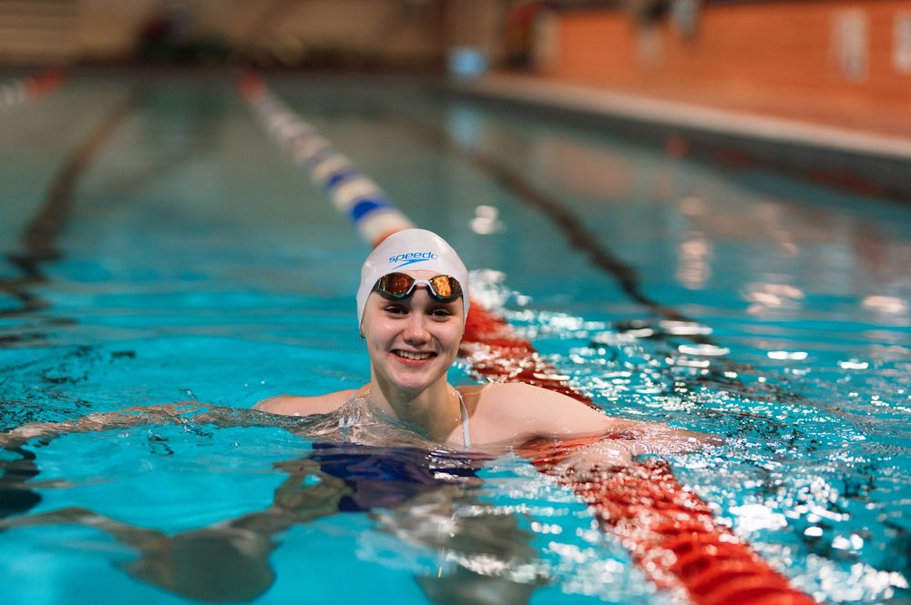 Competitive swimmer wearing goggles and white swim cap smiles while holding onto lane divider in indoor pool.