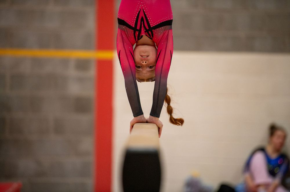 An upside-down gymnast performs on aerial silks with a red fabric against a white and gray interior background.