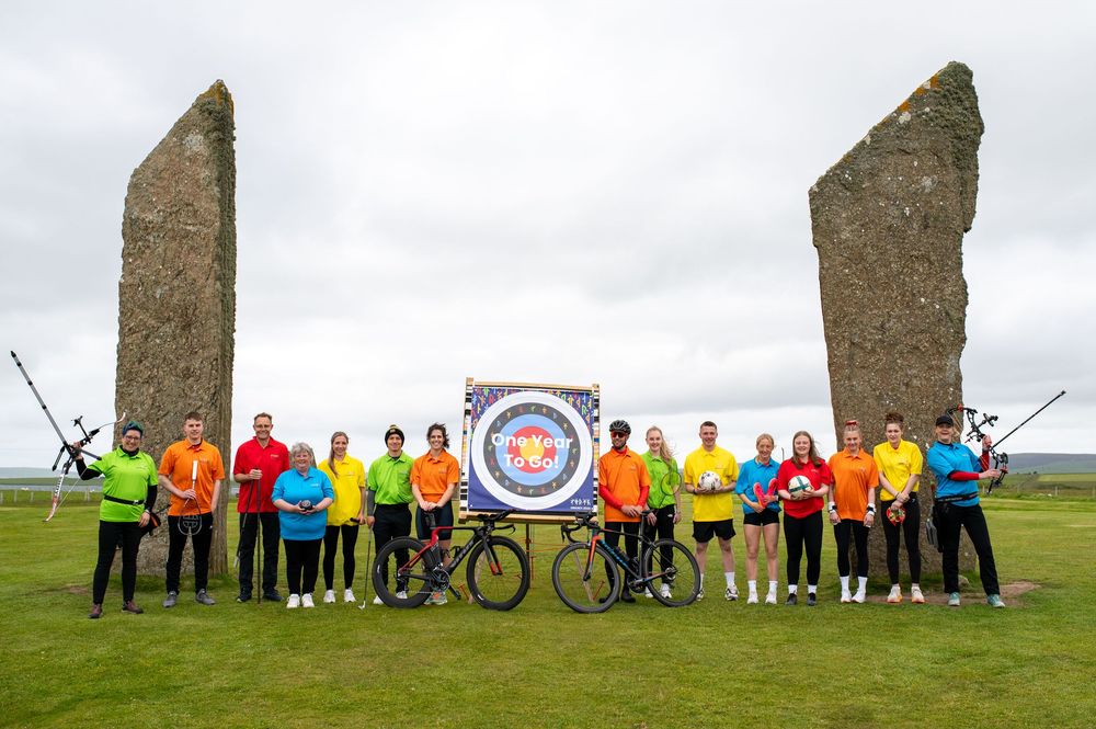 A group of cyclists in colorful jackets pose with their bikes between two ancient standing stones at an outdoor location.