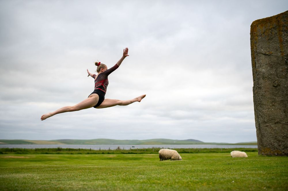 A dancer performs a graceful split leap on a green field near standing stones against a cloudy sky.
