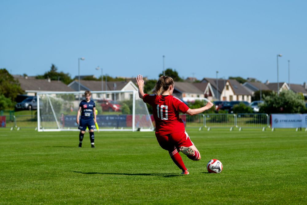 A soccer player in red dives to make a save while another player takes a shot on goal during an outdoor match.