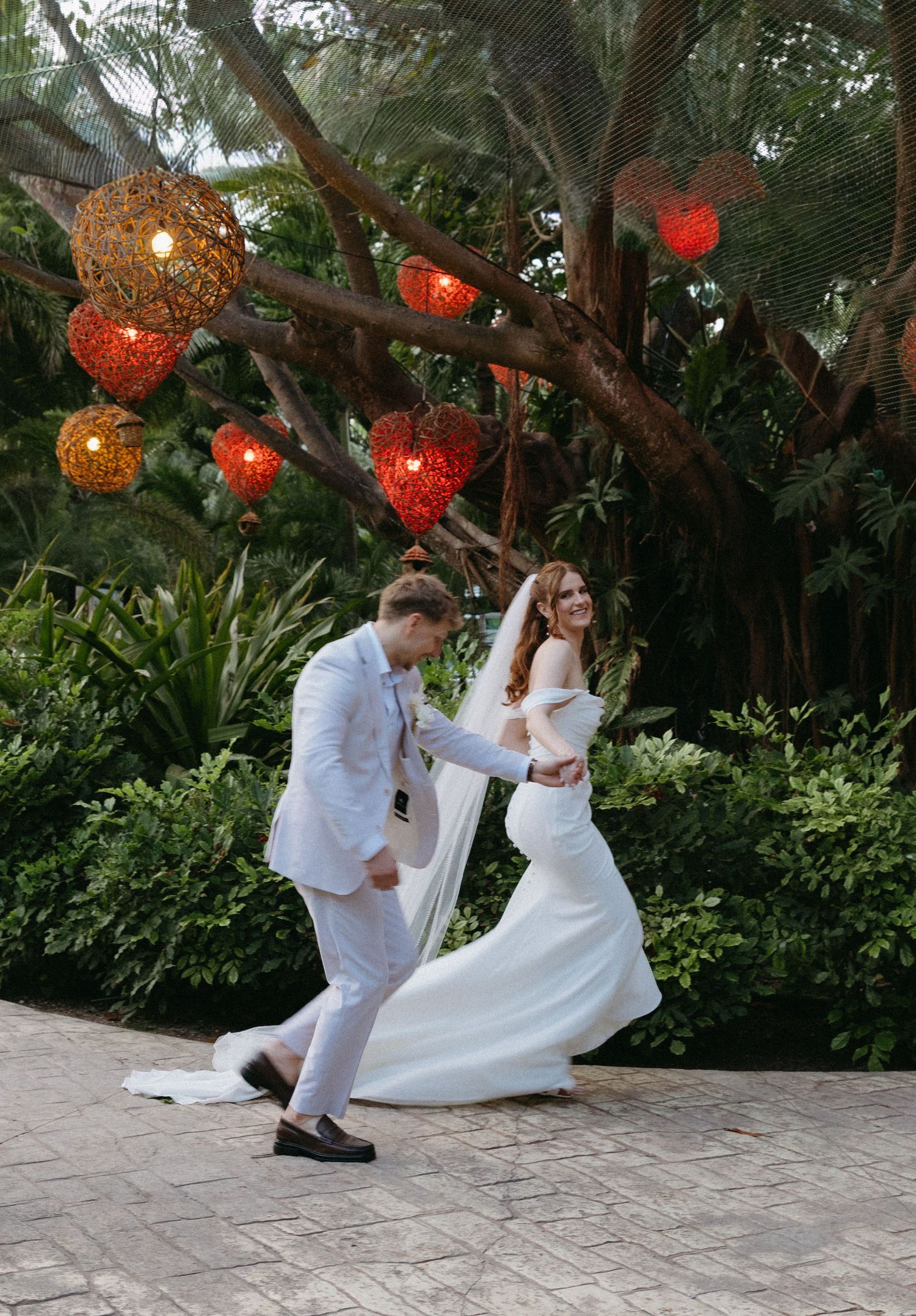 Joyful newlyweds dance under colorful hanging lanterns surrounded by lush tropical greenery at outdoor wedding.