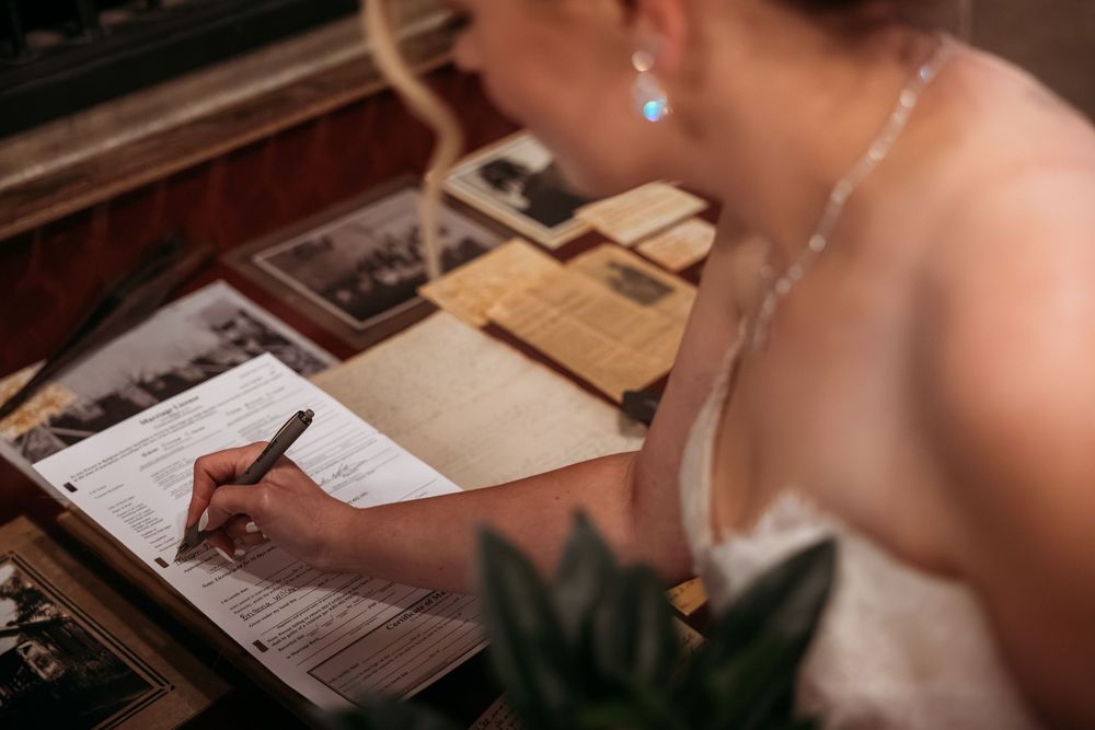 Bride in white dress signing wedding documents at a decorated desk with vintage photos and stationery.