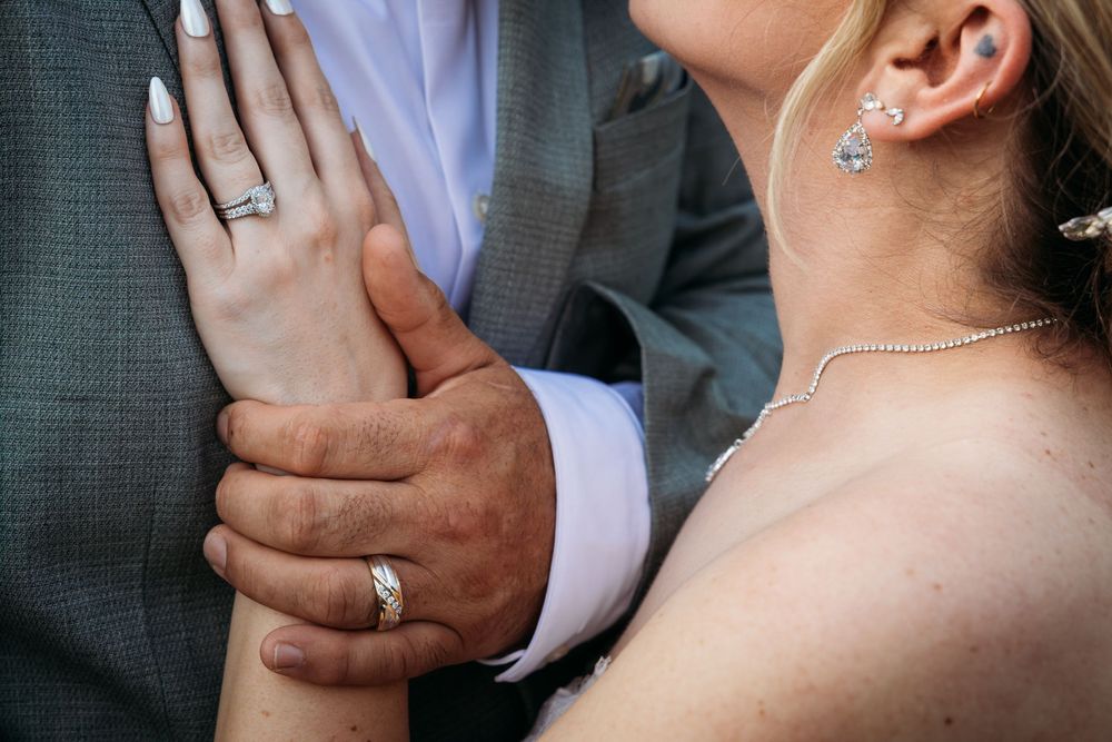 Close-up of a bride and groom holding hands, showcasing wedding rings, pearl necklace, and diamond earrings.