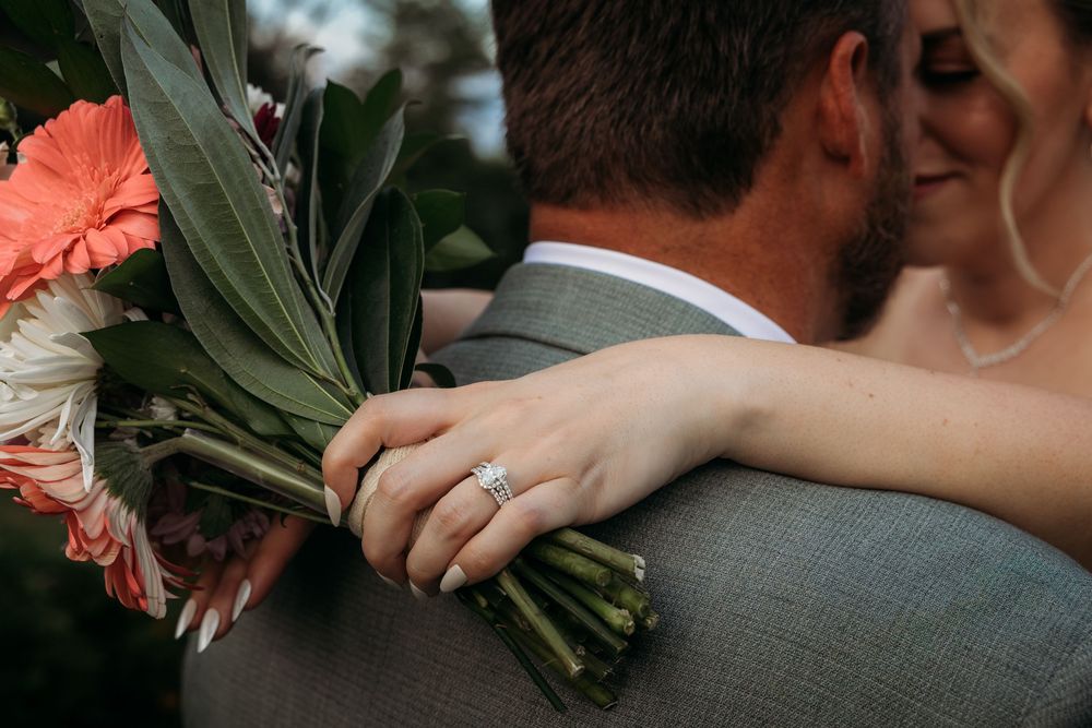 Bride rests hand with diamond engagement ring on groom's shoulder while holding coral wedding bouquet.