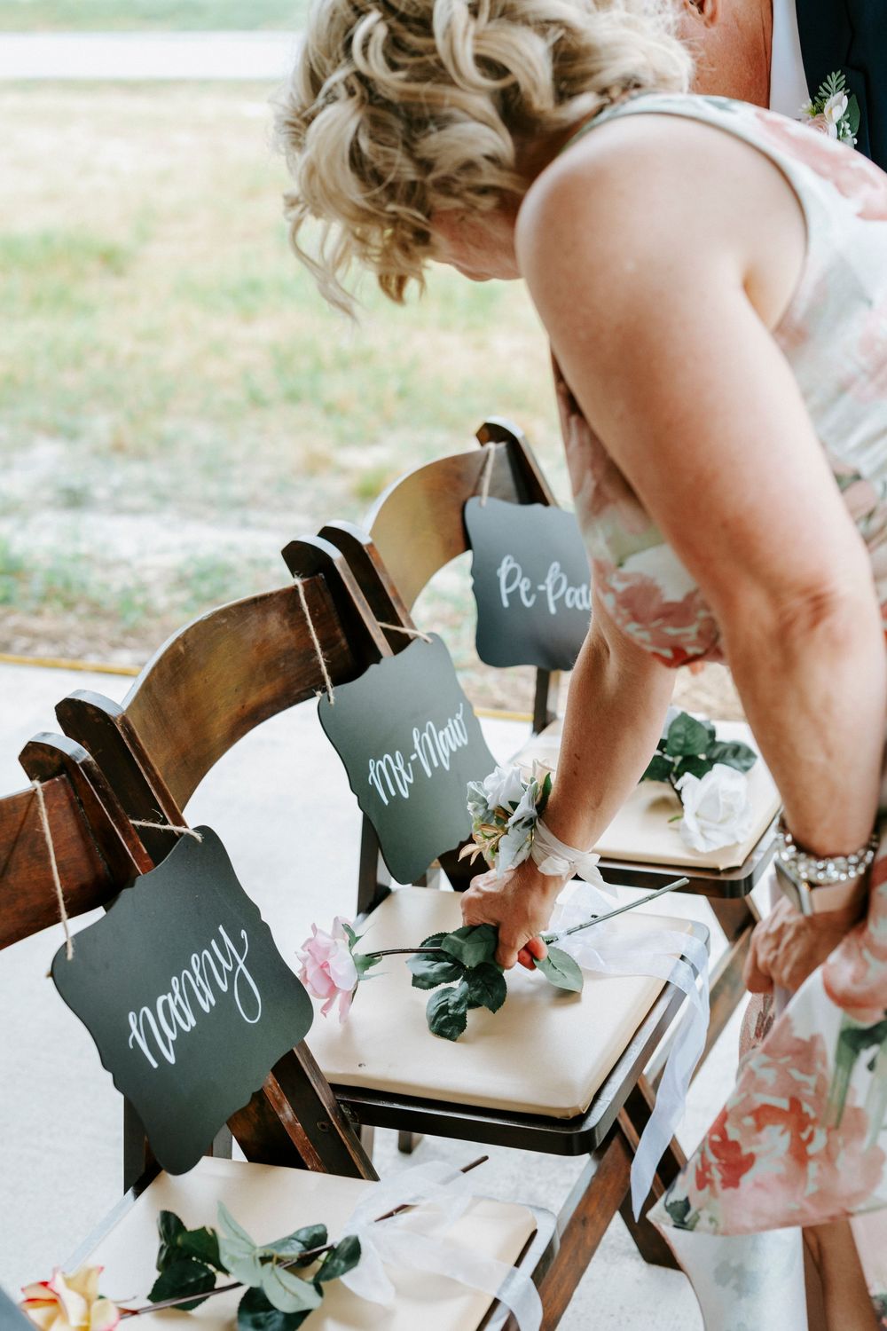Woman arranging floral decorations on wooden chairs with elegant calligraphy signs reading Nanny, Me-Maw, and Pe-Paw.