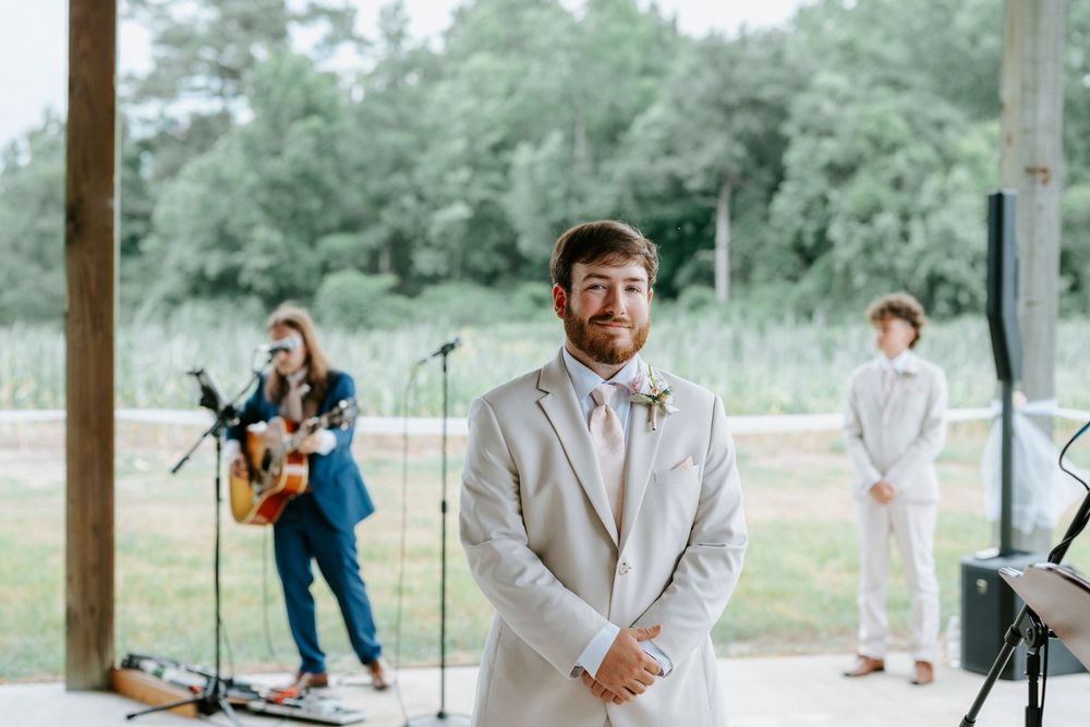 Groom in gray suit with pink tie stands at altar during outdoor wedding ceremony with guitarist performing in background.