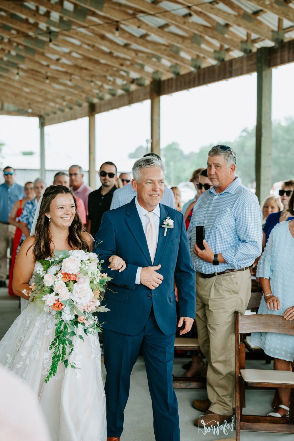 Bride walks down the aisle with her father in a blue suit at an outdoor wedding ceremony.