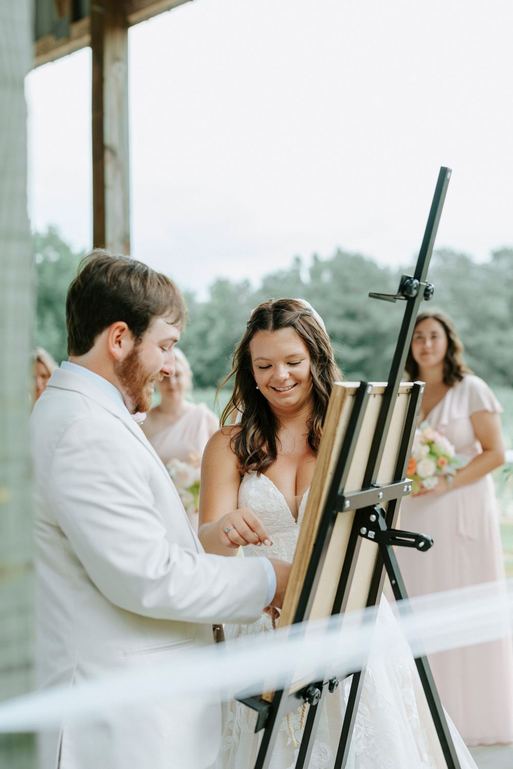 Bride and groom share a moment at an easel during their wedding ceremony with bridesmaids behind.