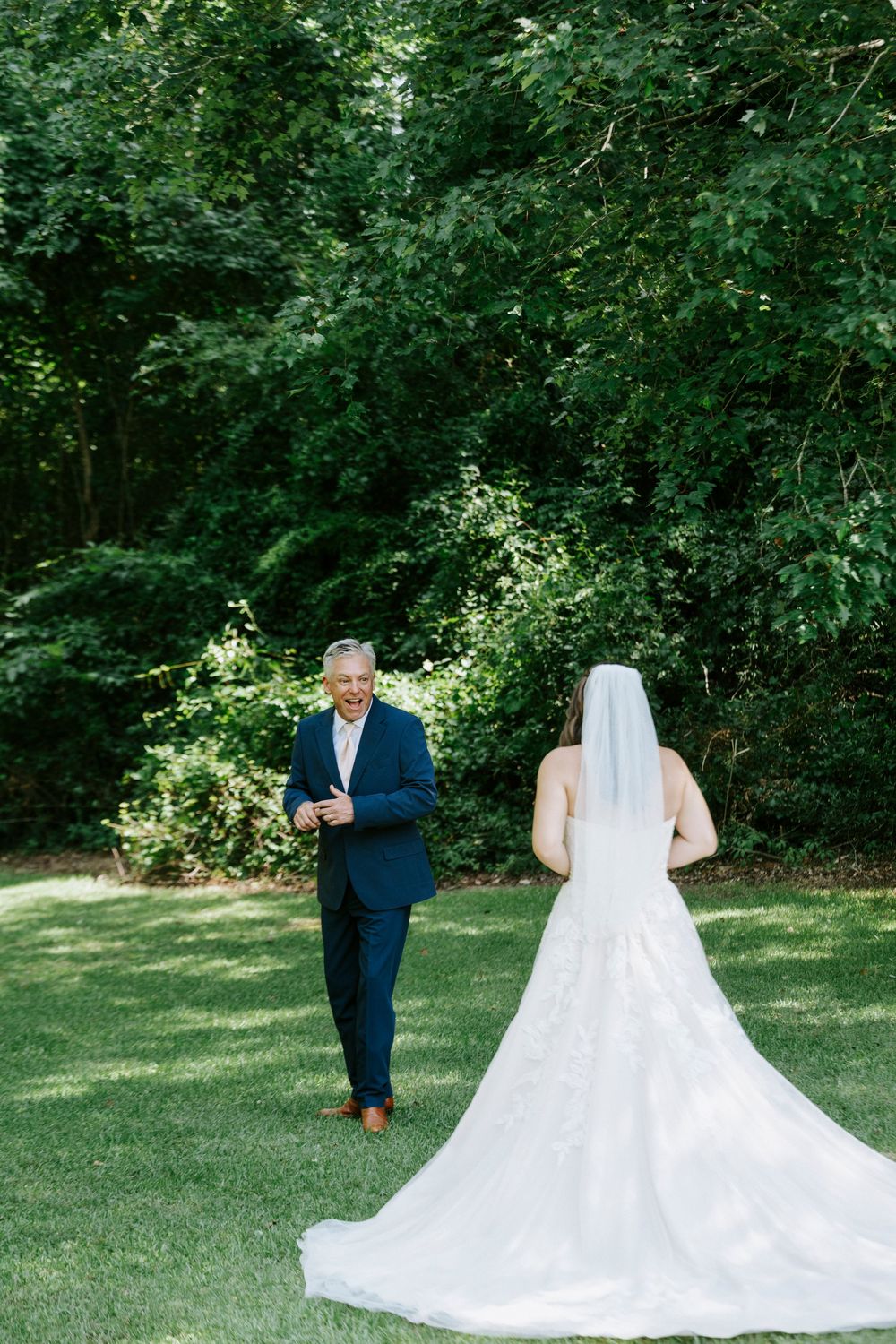 A bride in a white gown and veil shares a first look moment with a smiling man in a navy suit on a green lawn.