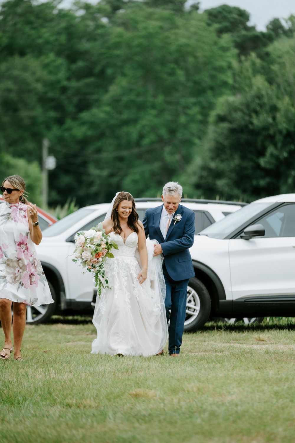 Bride in lace gown walks arm-in-arm with father across green lawn, carrying colorful bouquet at outdoor wedding.