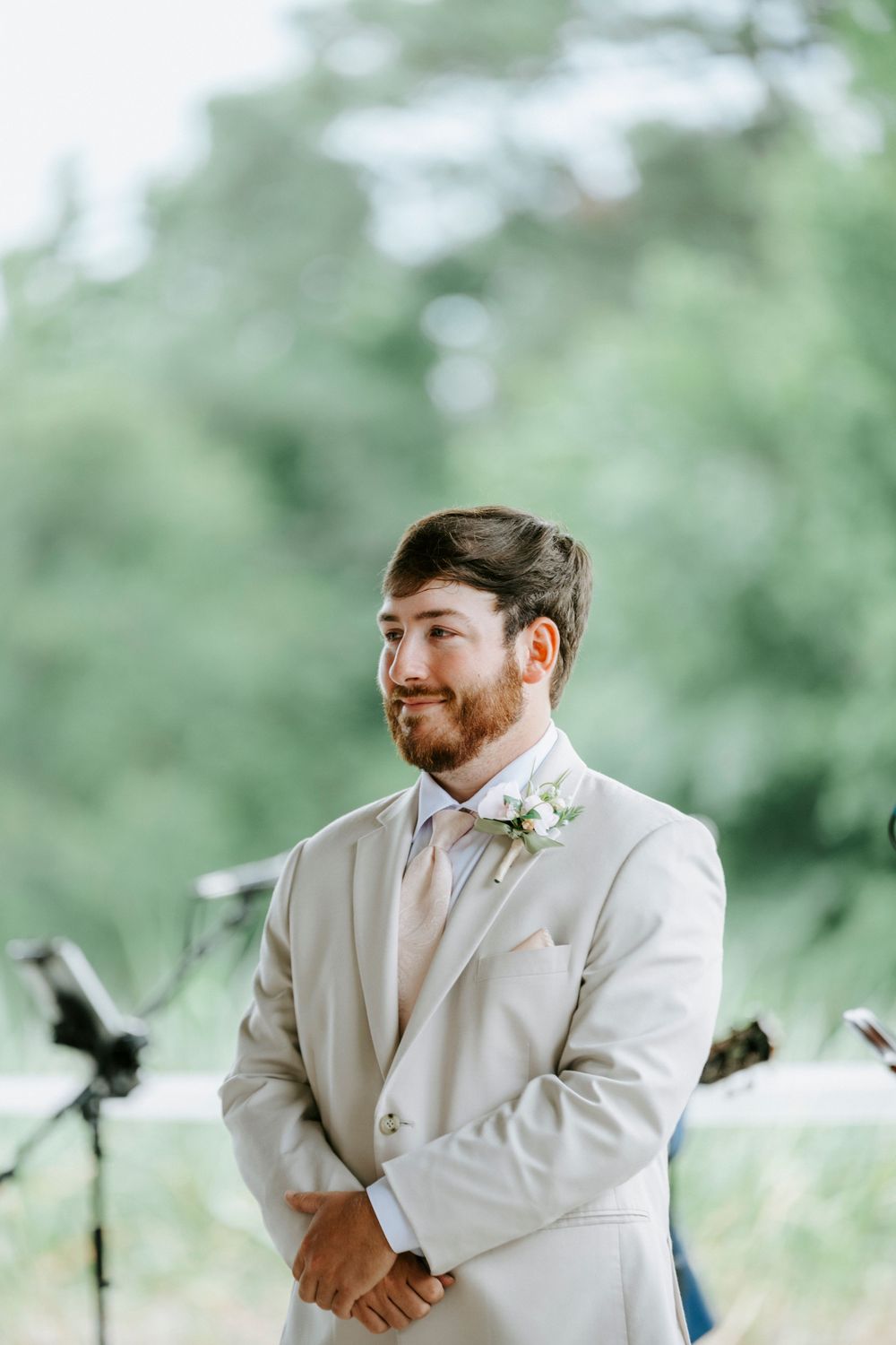 Groom in beige suit with boutonniere smiling during outdoor wedding ceremony.