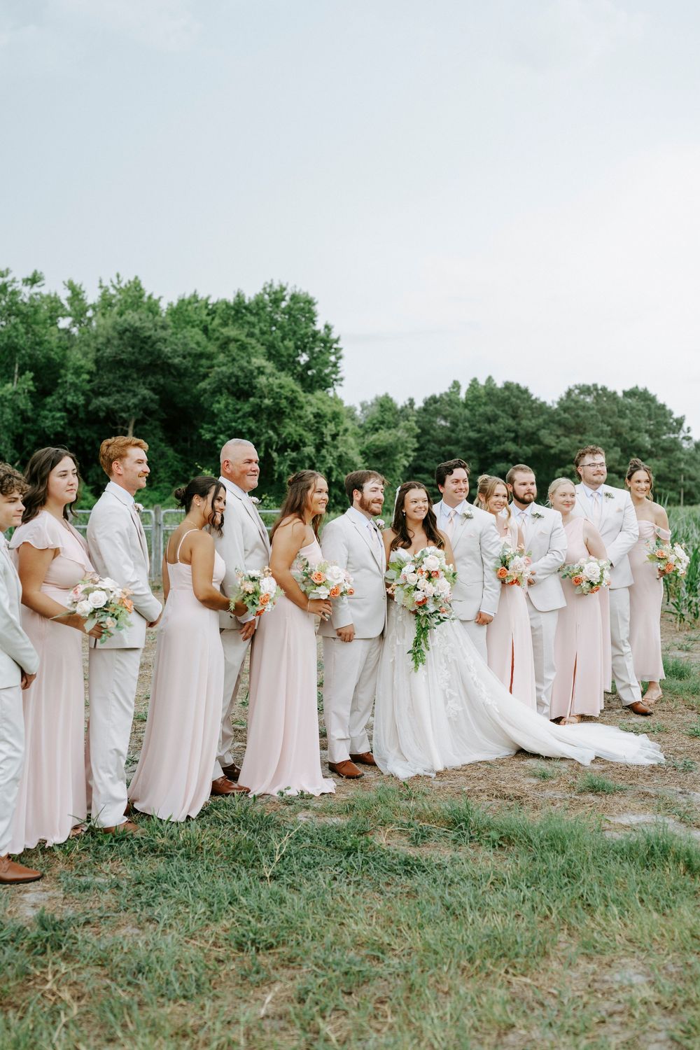 Wedding party poses outdoors, bridesmaids in blush gowns and groomsmen in tan suits flanking the bride and groom.