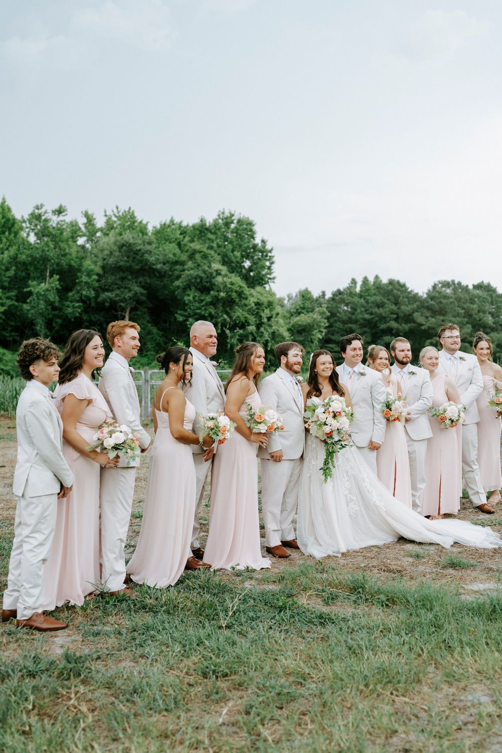 Bride and groom stand with their wedding party in blush and tan attire, laughing together in an outdoor setting.