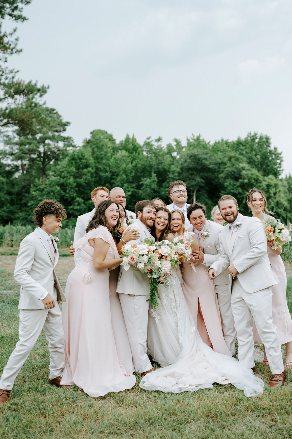 Joyful wedding party in blush and ivory attire laughing together outdoors surrounded by lush greenery.