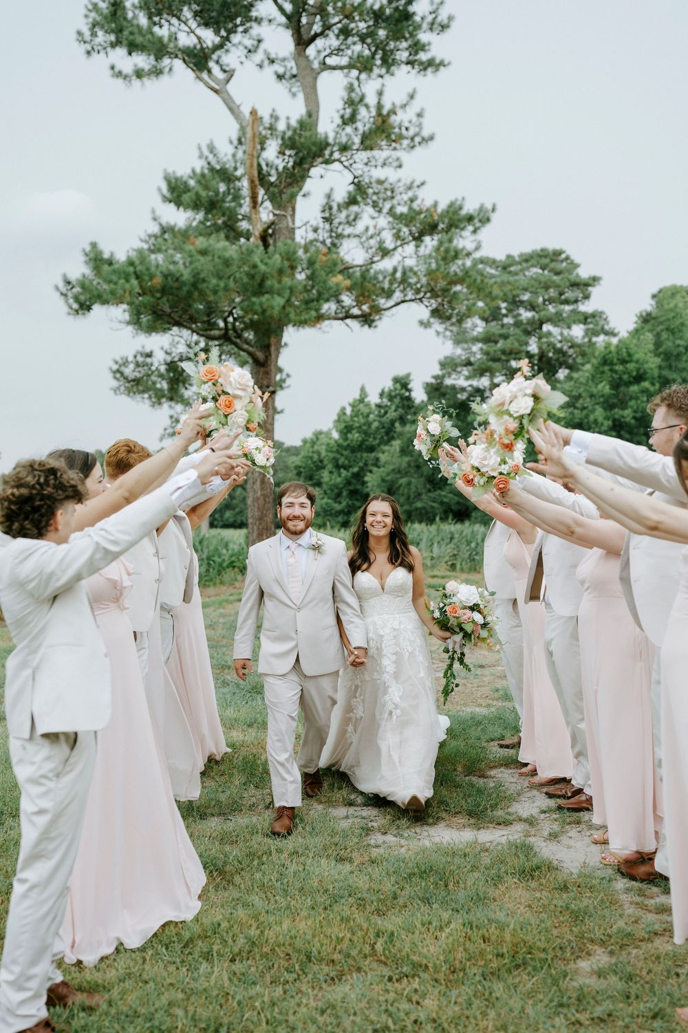 Bride and groom walk through bouquet arch with wedding party at outdoor ceremony.