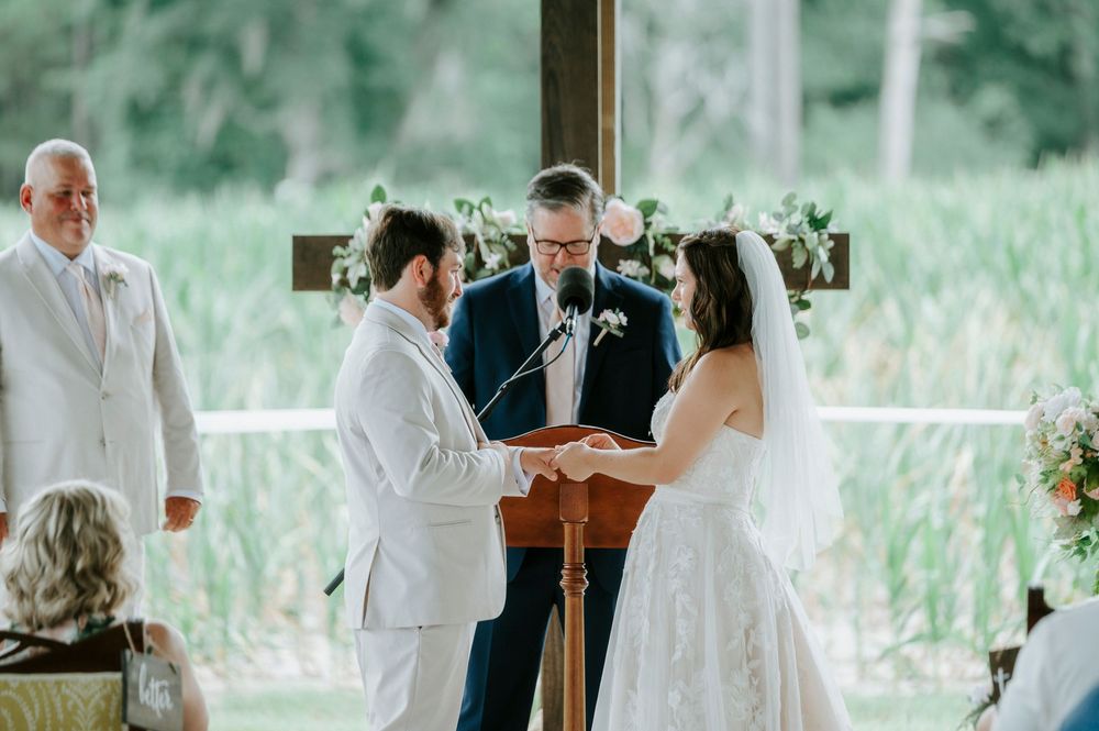 Bride and groom exchange rings at outdoor wedding ceremony with officiant at wooden podium.
