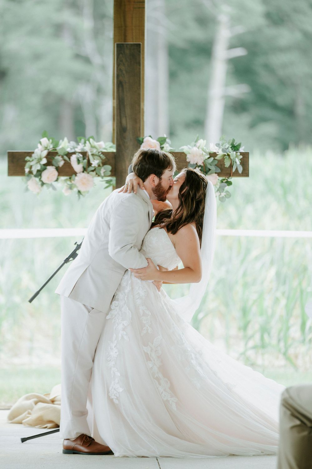 Bride and groom share their first kiss in front of a floral-adorned wooden cross at their wedding.