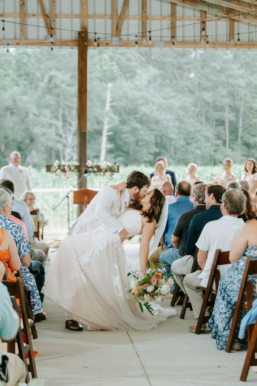 Bride and groom share romantic dip kiss during barn wedding ceremony with seated guests.