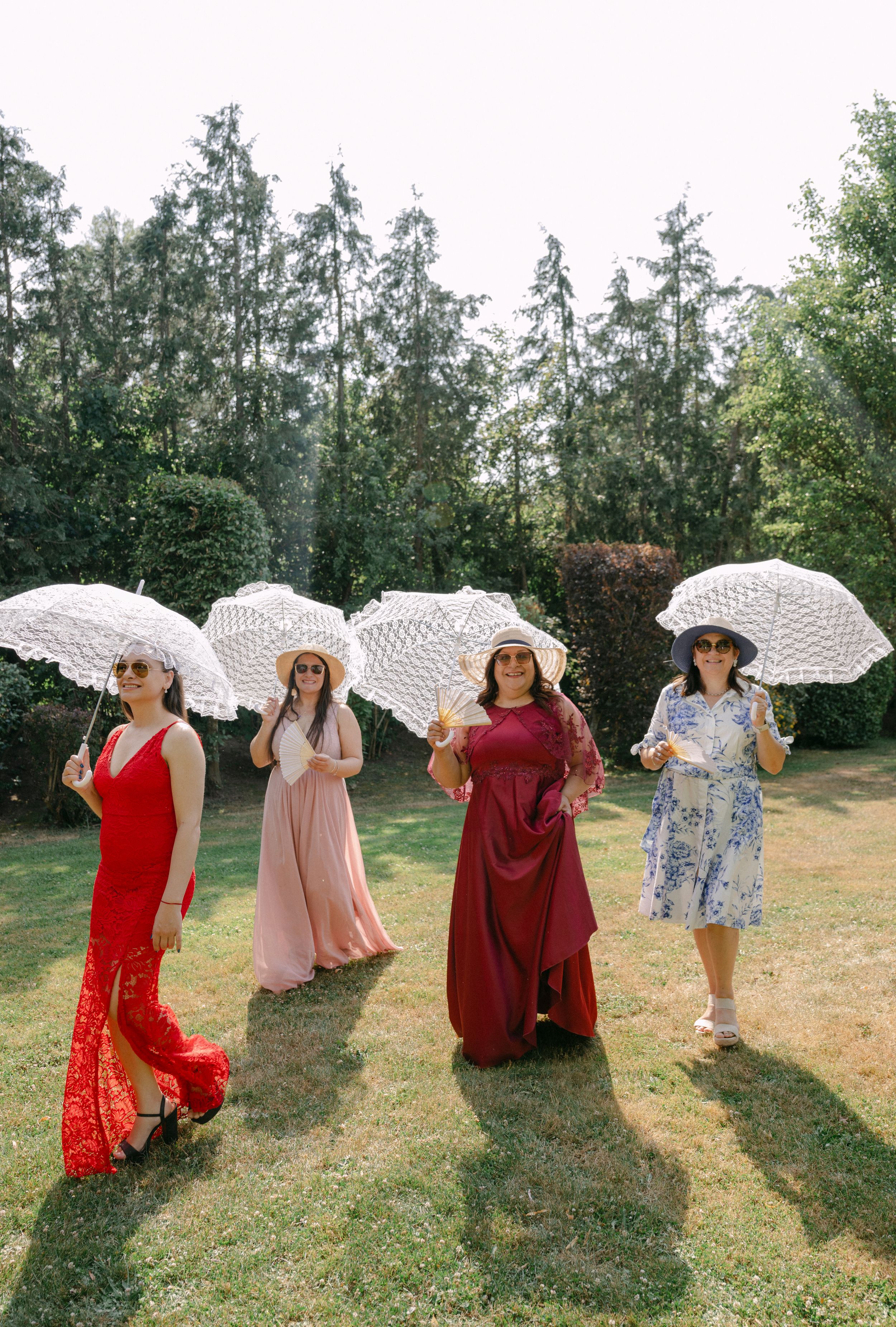 Four women in colorful dresses holding white lace parasols pose outdoors on a sunny day with lush green trees behind them.
