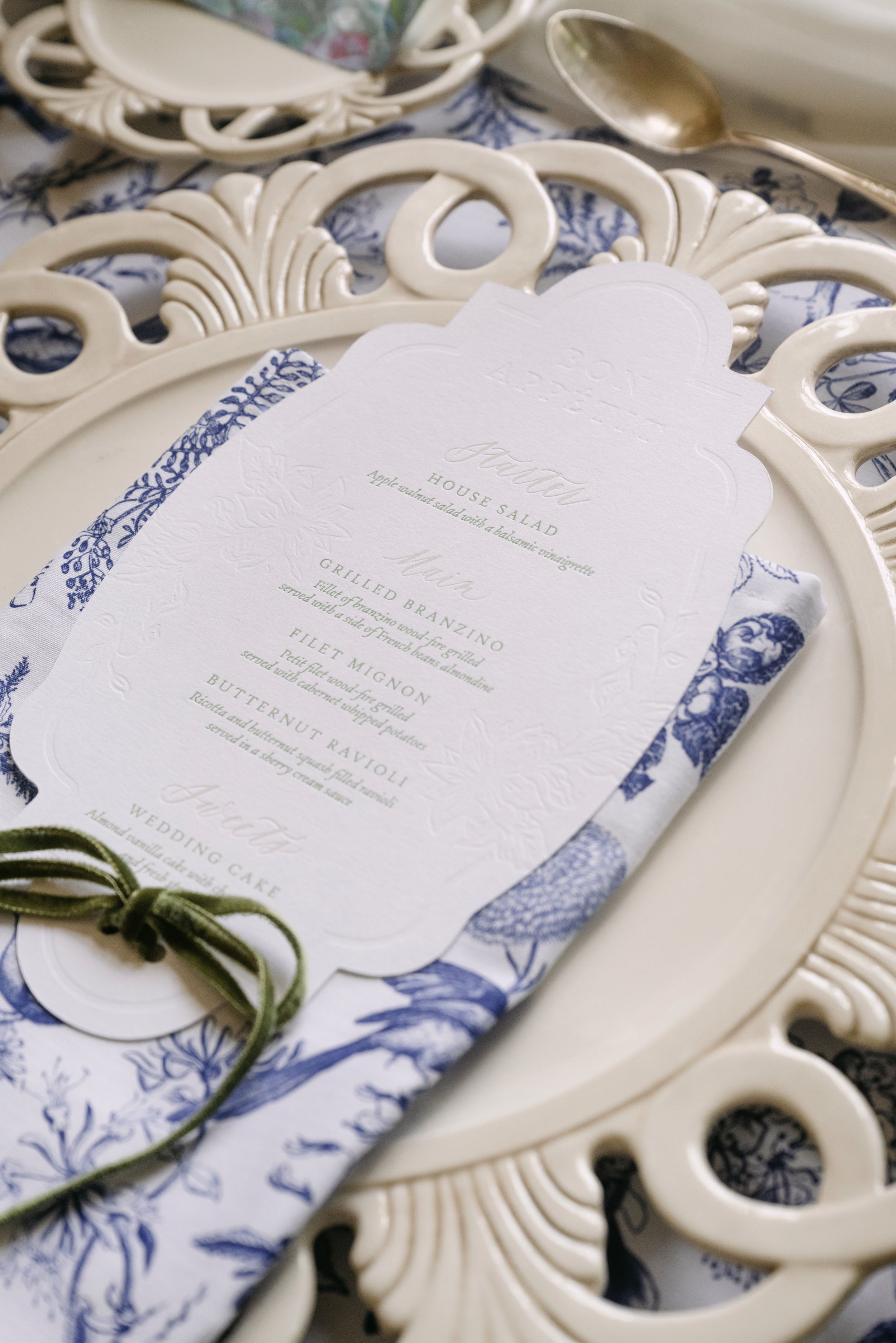 Elegant wedding place setting with blue toile napkin, ornate cream charger plate, and embossed menu card.