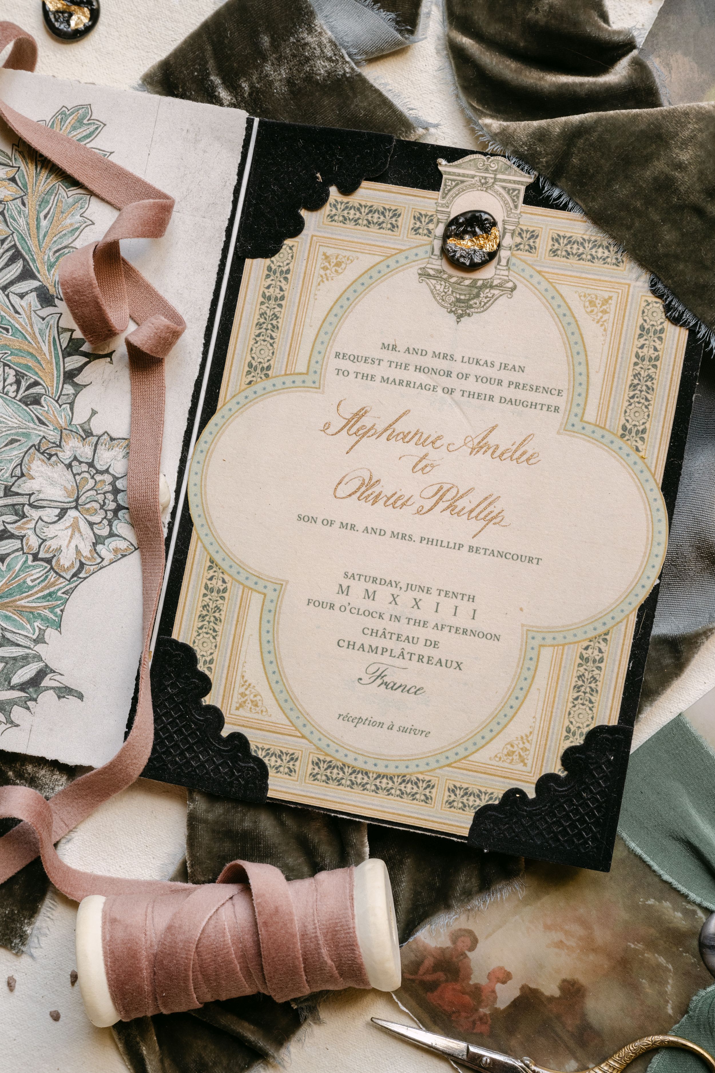 Elegant vintage wedding invitation with gold script, black lace border, and decorative brooch, surrounded by pink ribbons and floral paper.