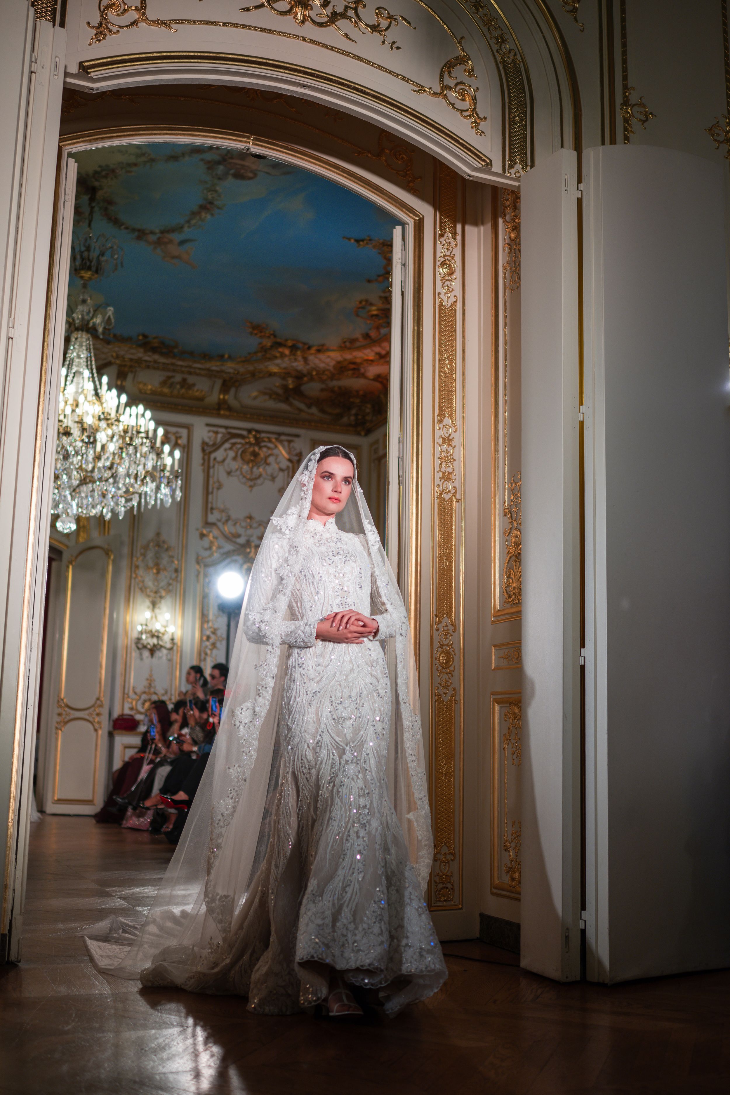 A model in an ornate ballroom wearing an embroidered white bridal gown with long veil and long sleeves.