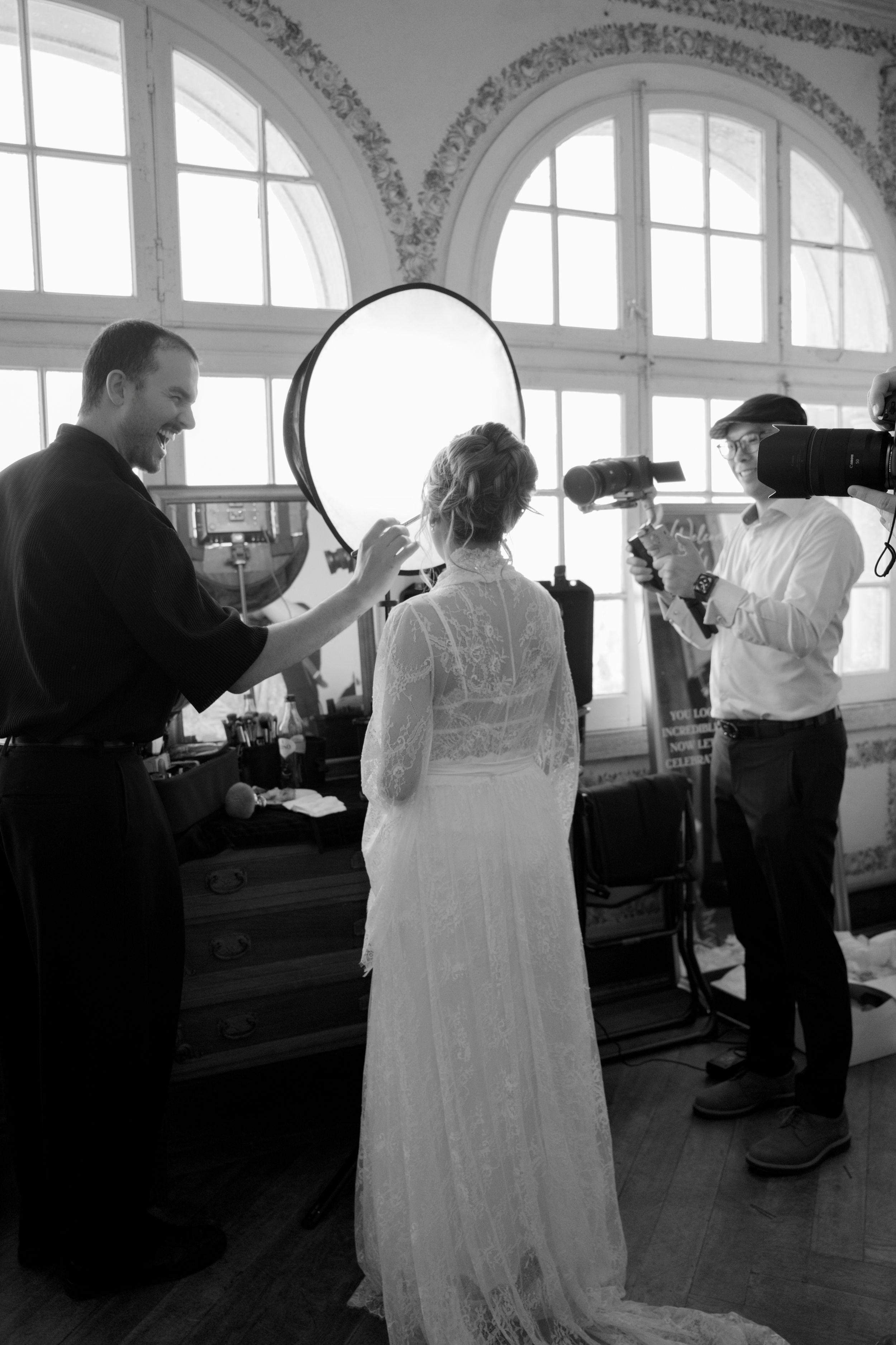 Black and white photo of a bride in lace gown getting makeup done while a videographer films the preparation.