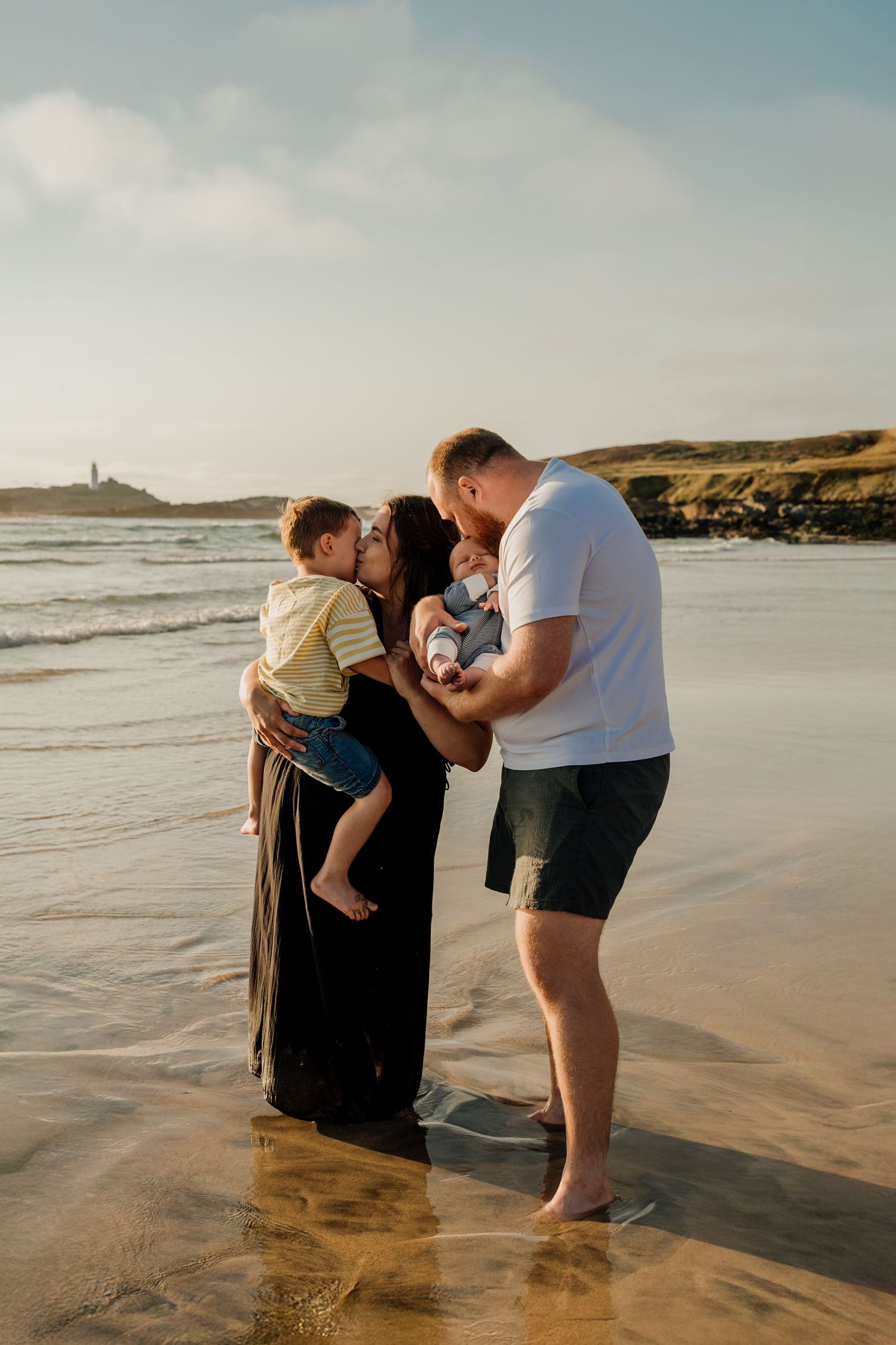 Family of four enjoying a golden hour beach photoshoot, parents kissing children by ocean waves.