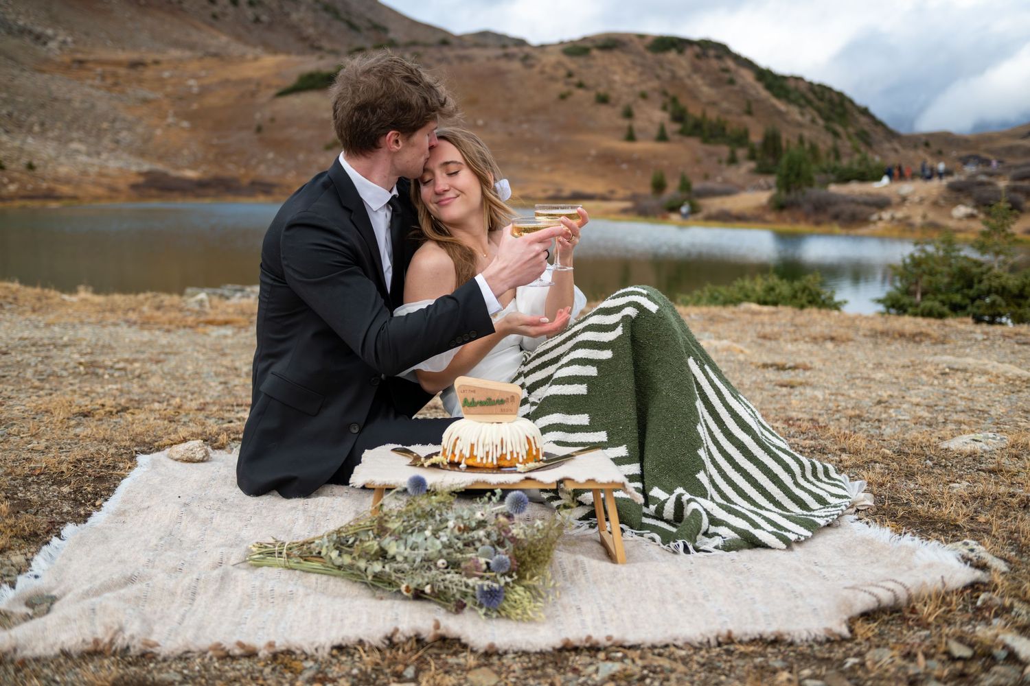 Couple shares a romantic lakeside picnic with cake and champagne, wrapped in a striped blanket amid mountain scenery.