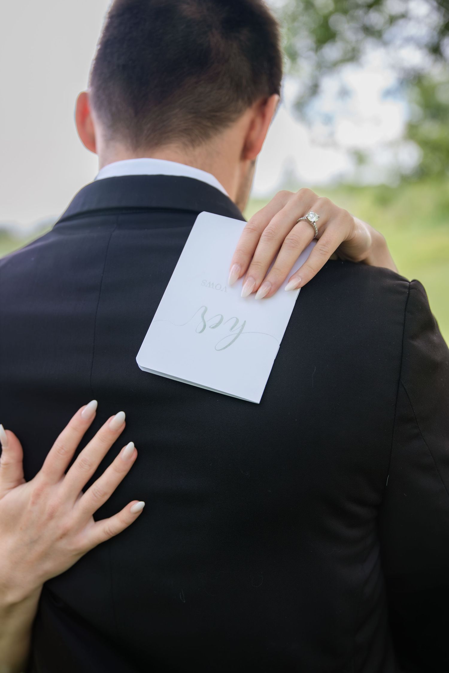Bride holding wedding vows card on groom's back in black suit, engagement ring visible, outdoor ceremony.