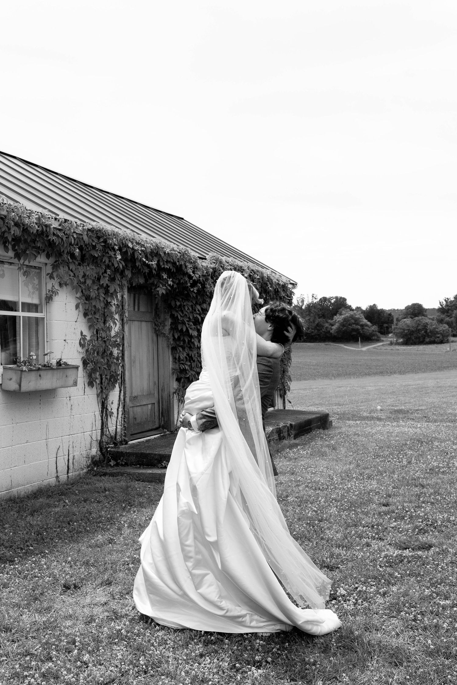 Black and white photo of a groom lifting his bride in a wedding dress outside a rustic ivy-covered building.