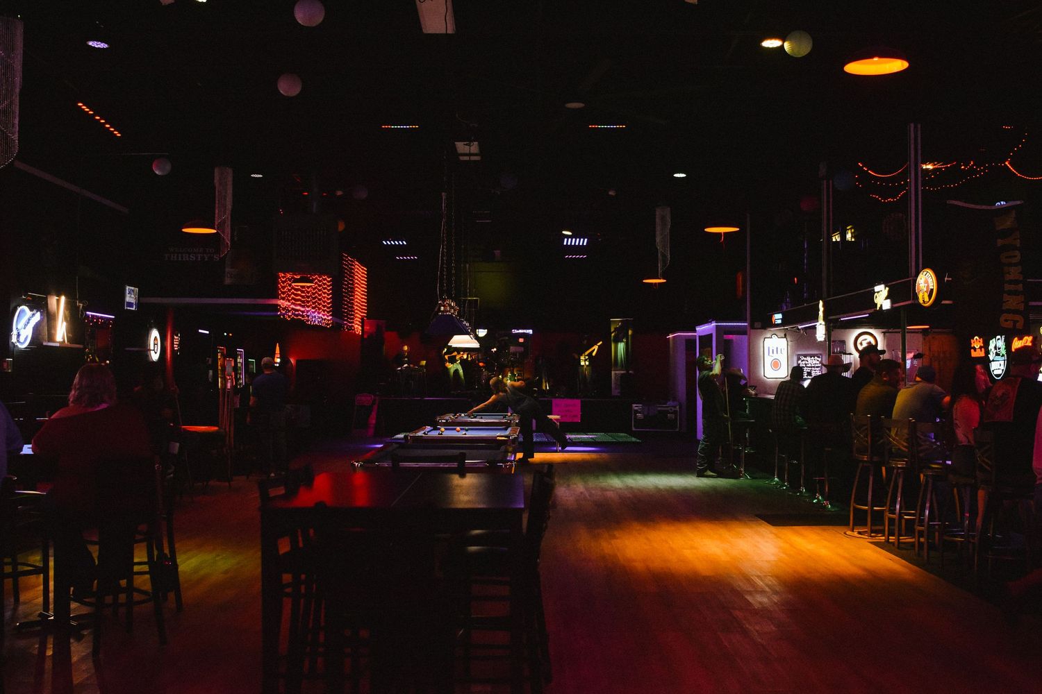 Dimly lit bar interior with pool tables, neon signs, and wooden dance floor in a nightclub setting.