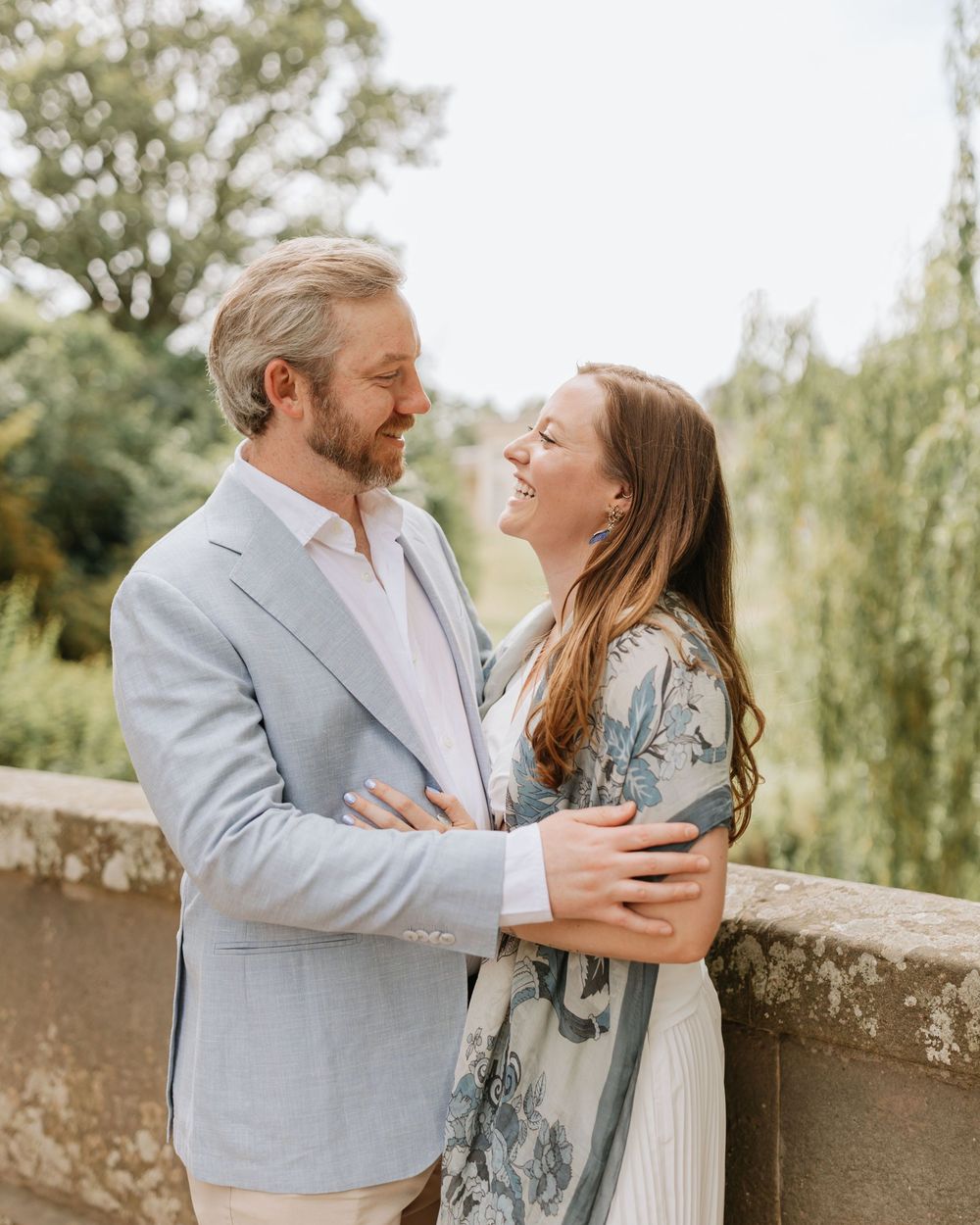 A couple embraces and smiles at each other outdoors, with lush greenery and a stone wall in the background.