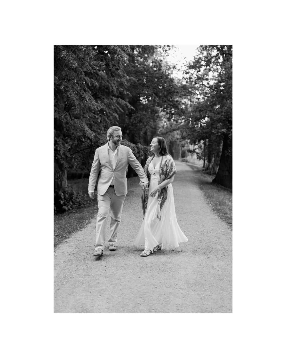 Black and white photo of couple walking hand in hand down a tree-lined garden path.