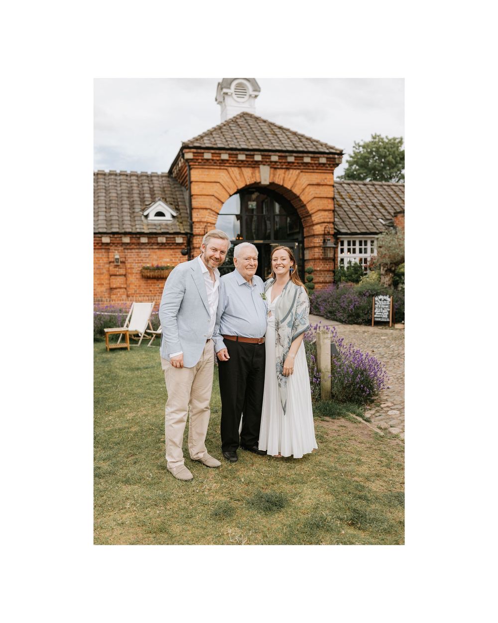 Three people smiling together outdoors in front of a brick archway building surrounded by lavender gardens.