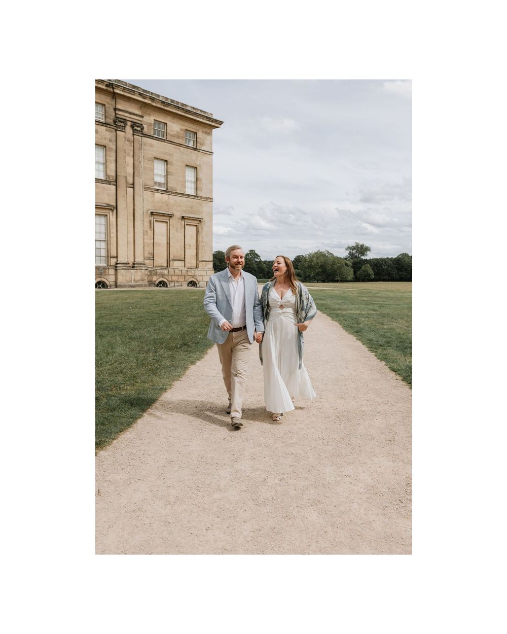 Elegant couple walking hand in hand on a gravel path beside a grand estate building.