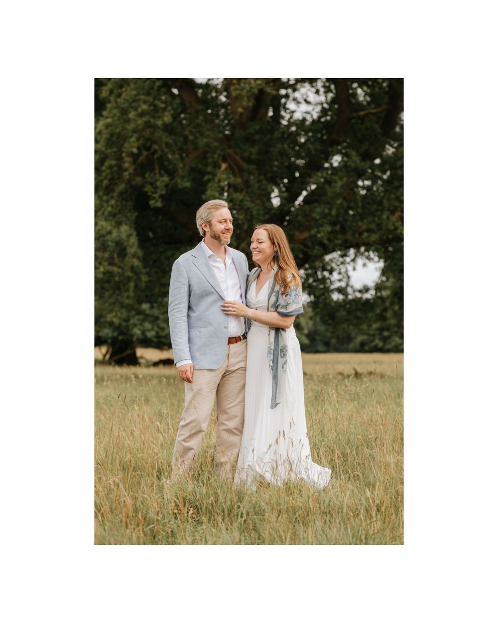 A couple stands in a lush meadow, the woman in a white gown and the man in a light blue blazer.