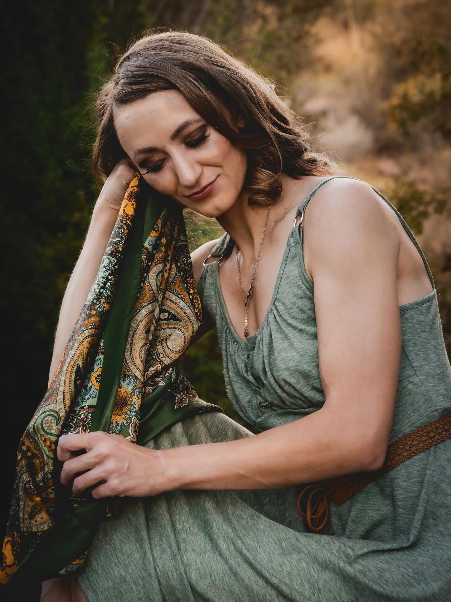 A woman in a sage green dress holds a paisley scarf, posing outdoors with a serene expression.