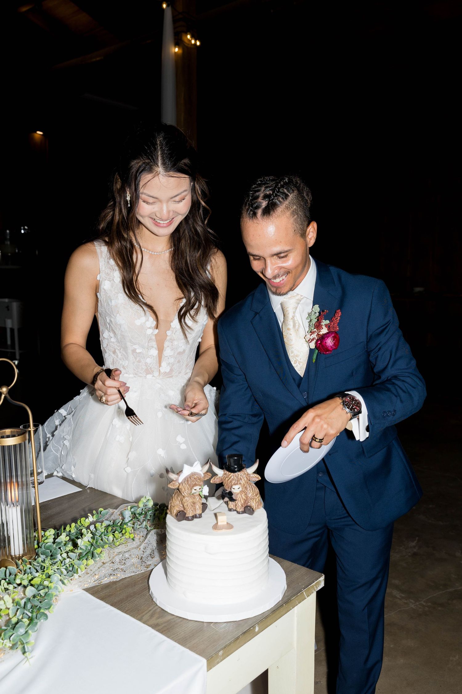Bride and groom cutting white tiered wedding cake together, bride in lace gown, groom in navy suit with boutonniere.
