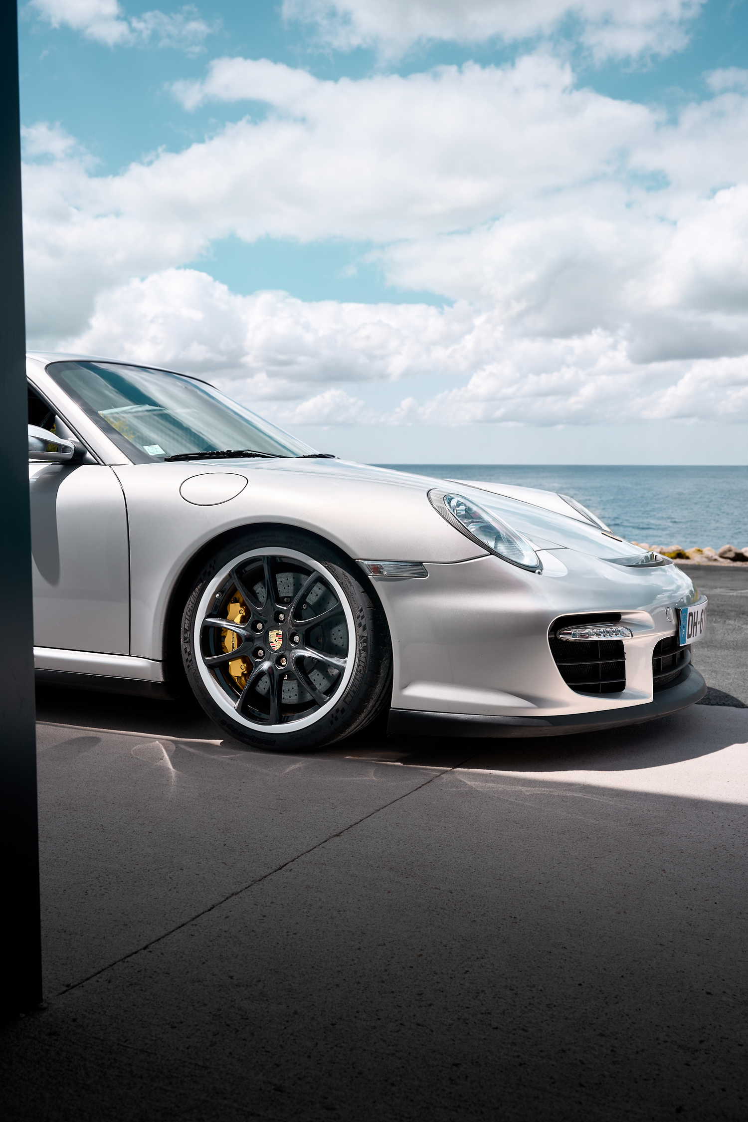 Silver Porsche 911 parked by the ocean under a partly cloudy sky, showcasing its sleek front end and black alloy wheels.