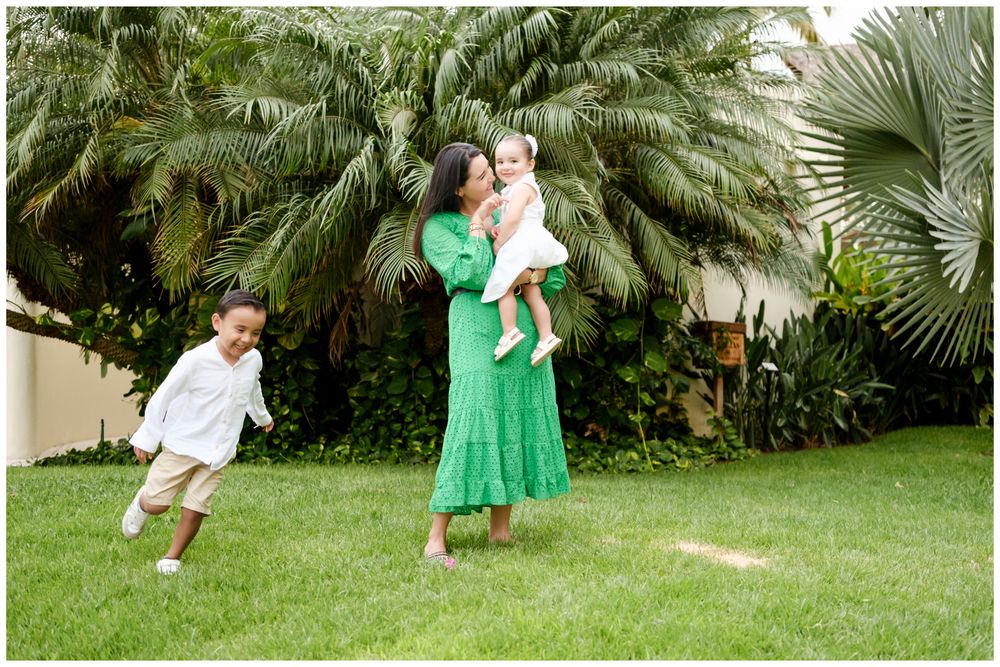 Mother lifting her baby on a Punta Mita lawn while her child runs nearby, candid family photography Riviera Nayarit.