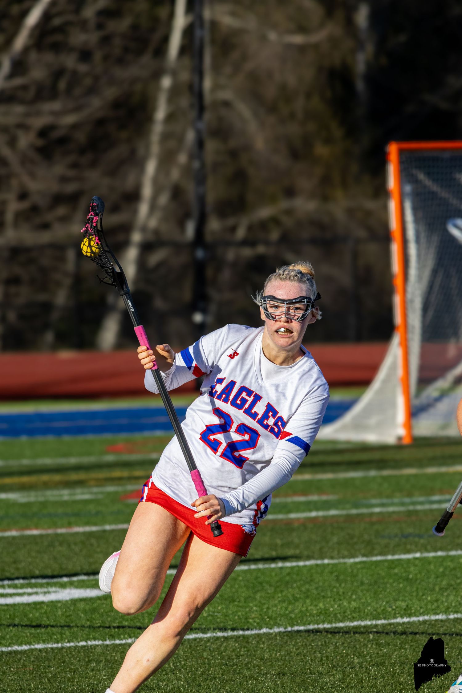 Female lacrosse player #22 in Eagles uniform sprints with stick during outdoor game on turf field.
