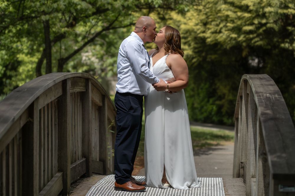 Couple sharing a romantic kiss on a wooden bridge surrounded by lush green trees in an outdoor wedding setting.